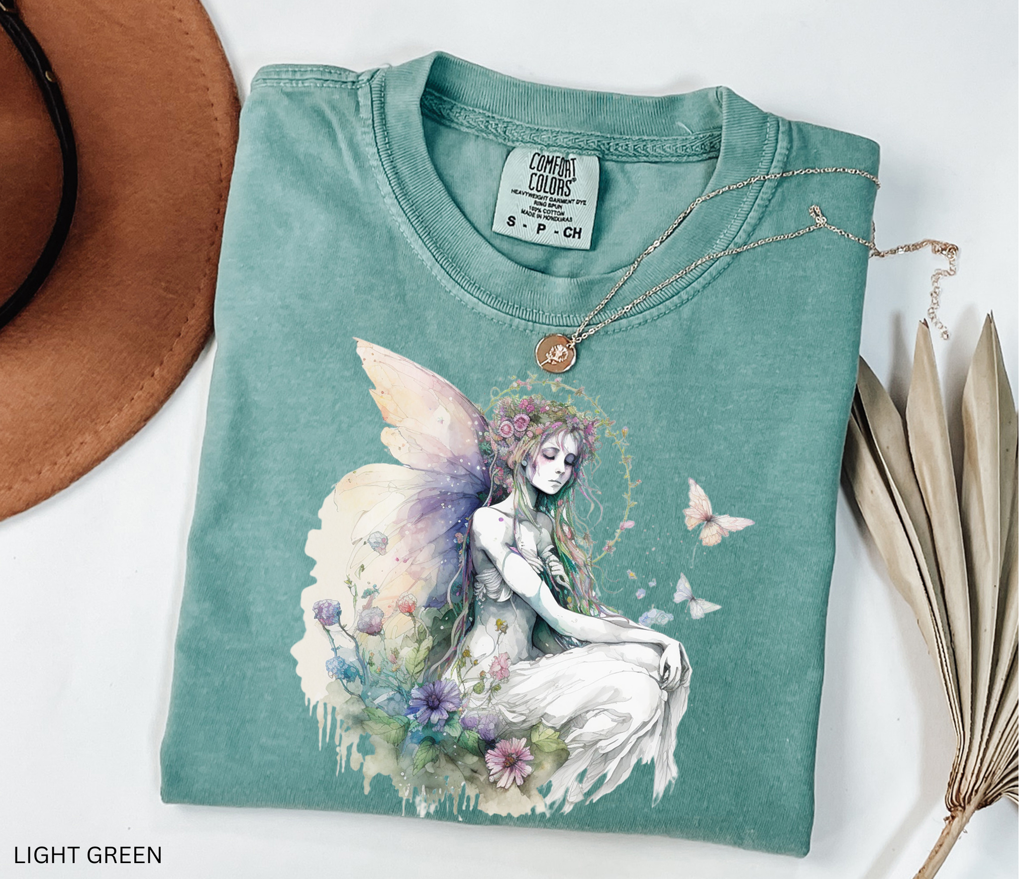 A folded light green t-shirt with a detailed fantasy fairy graphic, styled on a white background with a brown hat, dried palm leaves, and a gold necklace.