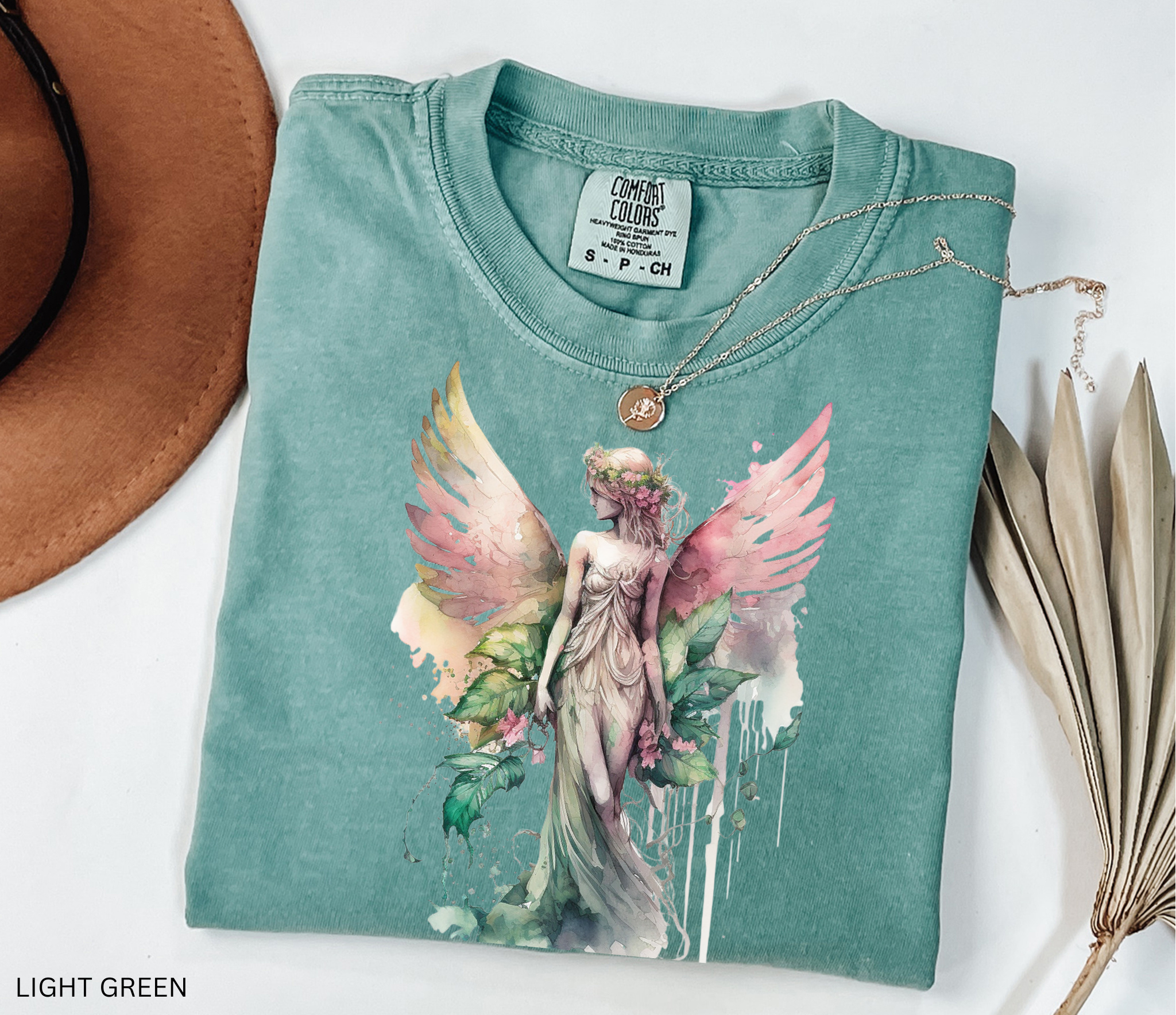 A folded light green graphic T-shirt with a watercolor-style angel design in pink, green, and cream tones, styled on a white background with a brown hat edge, dried leaves, and a necklace.
