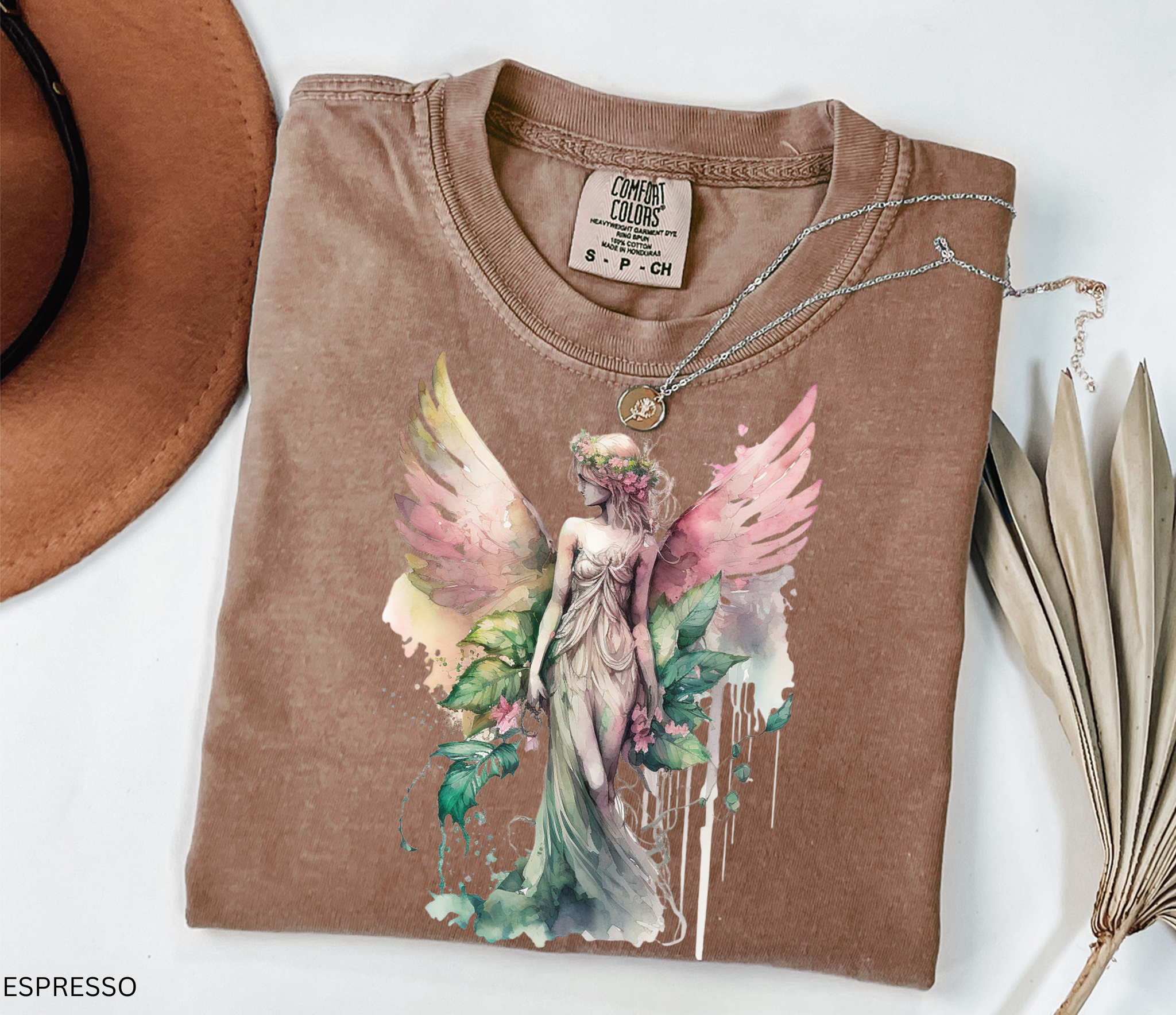 A folded espresso brown t-shirt featuring a colorful graphic of a winged fairy, styled on a white background with a brown hat, a silver necklace, and dried palm leaves.