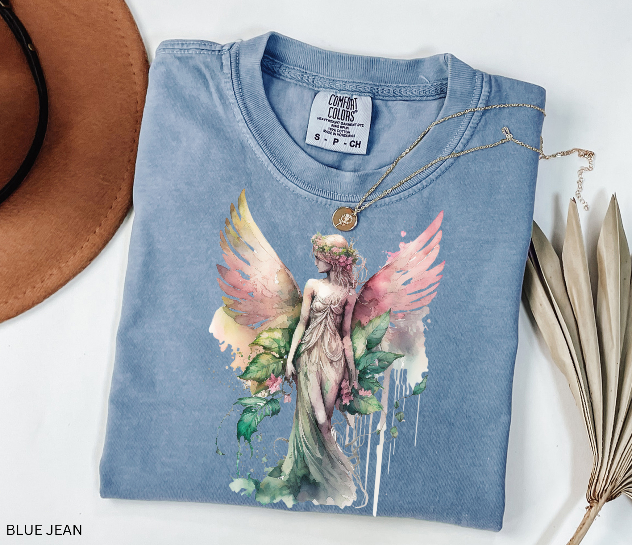 A folded light blue t-shirt with a graphic of a winged fairy surrounded by leaves, styled on a white background with a brown hat corner, a gold necklace, and dried leaves. The text 'BLUE JEAN' is visible in the bottom-left corner.