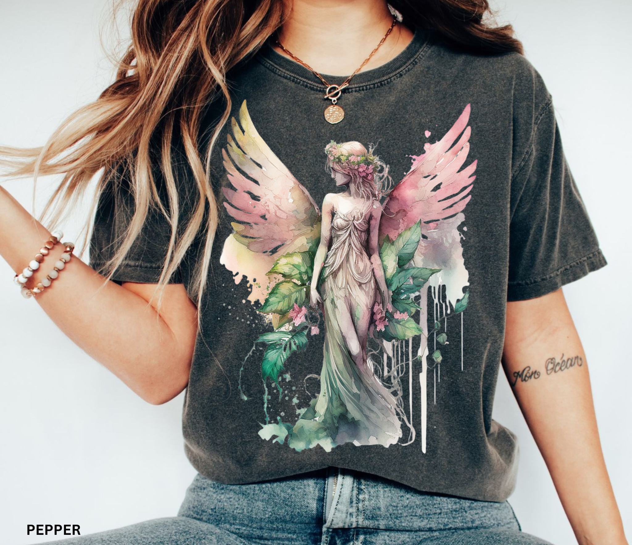 A woman wearing a dark charcoal gray graphic t-shirt with a colorful print of a winged goddess surrounded by green leaves and pink flowers. The text 'PEPPER' is visible in the bottom-left corner.