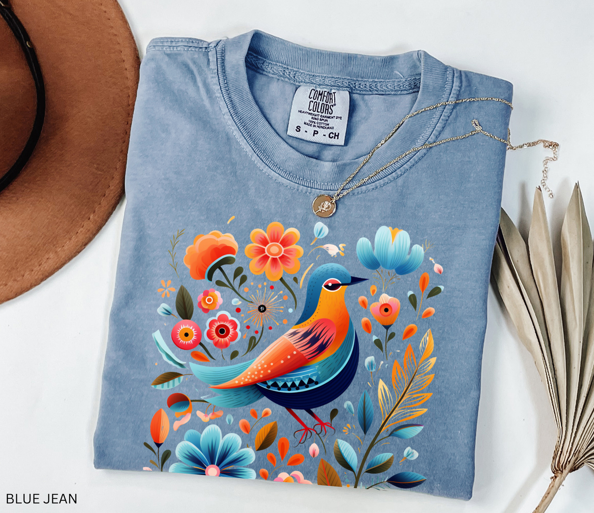 A folded light blue t-shirt with a colorful bird and floral graphic print, styled on a white background with a brown hat corner, dried leaves, and a necklace.