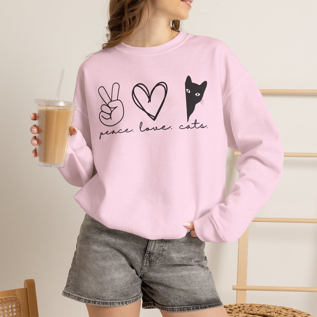 Woman holding an iced coffee while wearing a pink Peace Love Cats crewneck sweatshirt featuring a peace sign, heart, and black cat graphic.