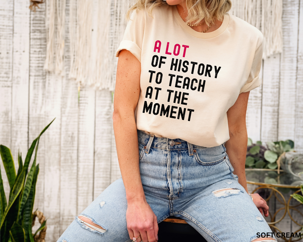 A woman wearing a cream-colored graphic t-shirt with the text 'A LOT OF HISTORY TO TEACH AT THE MOMENT' in pink and black lettering, paired with distressed light-wash blue jeans, posed in front of a rustic white wood wall.