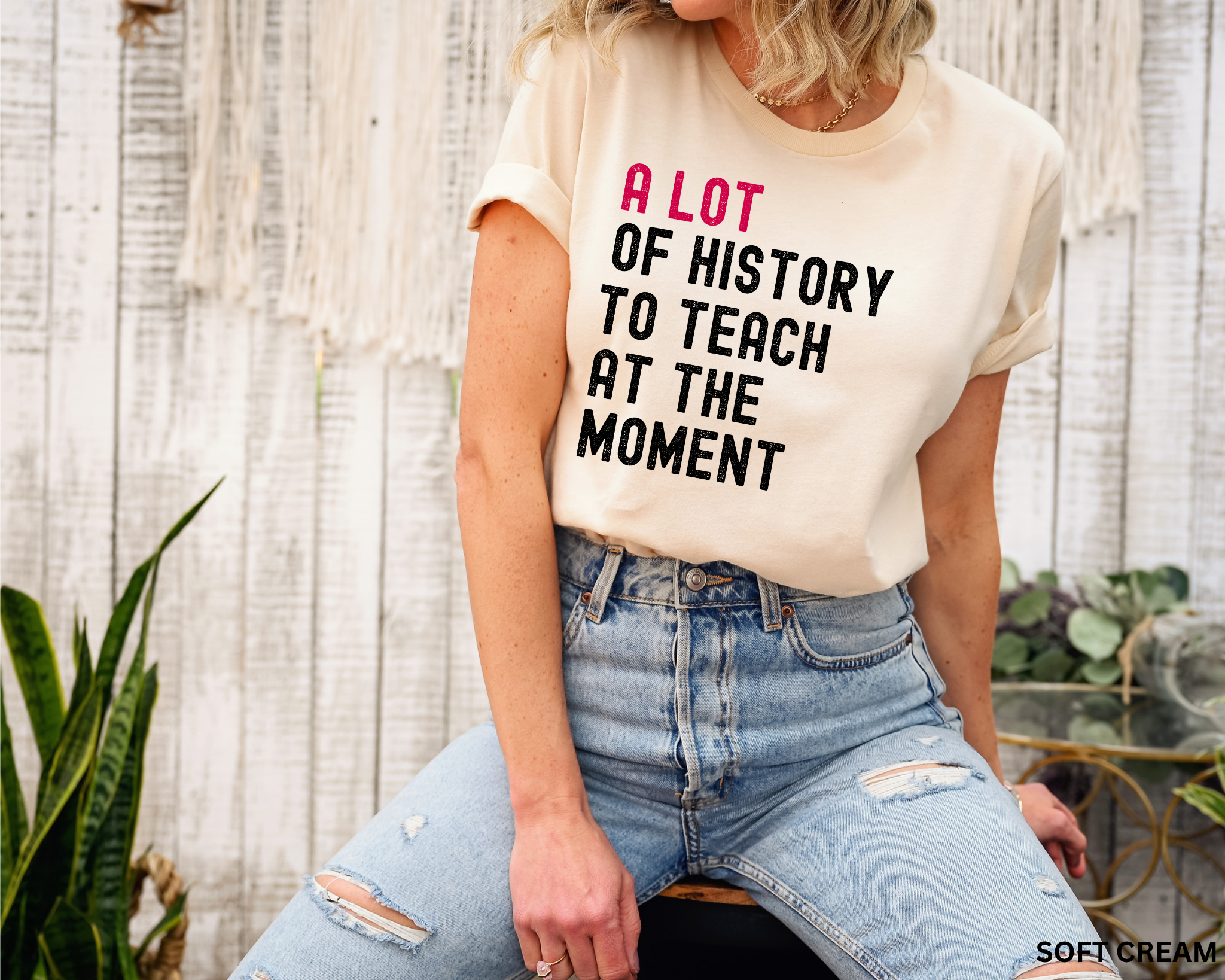 A woman wearing a cream-colored graphic t-shirt with the text 'A LOT OF HISTORY TO TEACH AT THE MOMENT' in pink and black lettering, paired with distressed light-wash blue jeans, posed in front of a rustic white wood wall.